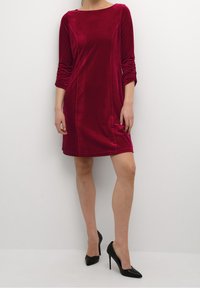 Röd sammetklänning med trekvartsärmar, en båturringning och rak passform. I kombination med svarta spetsskor med hög klack. Enkel, slät textur.