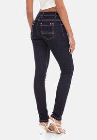 Dunkelblaue, figurbetonte Jeans mit hoher Taille, kontrastierenden Nähten und zwei Gesäßtaschen. Das Outfit wird mit weißen, schmalen High Heels kombiniert.