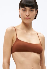 Sieviete ar īsiem tumšiem matiem un zelta riņķveida auskariem, kura valkā rūsas krāsas plānas lences bikini augšdaļu uz vienkārša gaiša fona.