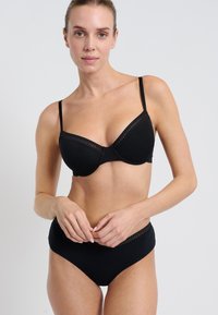 Woman wearing black bra and matching high-waist panties with lace trim, standing against a plain light background, hands clasped in front.