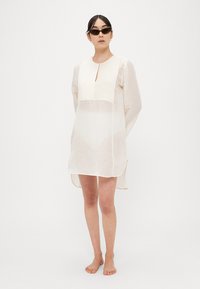 Abito trasparente color avorio chiaro con corpetto testurizzato, scollo a goccia e maniche lunghe, caratterizzato da una vestibilità comoda e spacchi laterali per facilitare i movimenti.