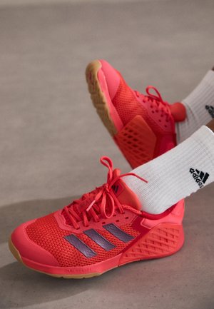 Stopy noszące jaskrawoczerwone sportowe buty Adidas z białymi skarpetkami z logo Adidas na szarym tle.