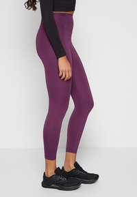 Legginsy z wysokim stanem, dopasowane, w głębokim fioletowym kolorze, wykonane z elastycznej tkaniny. W połączeniu z czarnymi butami sportowymi i czarną bluzą z długim rękawem.