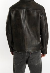 Musta nahkatakki mattapintaisella viimeistelyllä, yksityiskohtaiset hihansuut ja räätälöity istuvuus, yhdistetty harmaiden farkkujen kanssa.