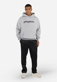 Gray hooded sweatshirt with “PEGA-DOR” printed in dark script, paired with black pants featuring a subtle logo and side stripes. Cream sneakers.