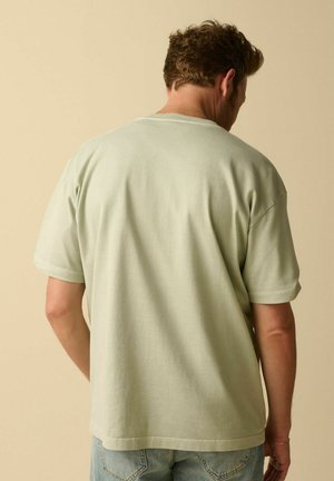 Homme aux cheveux courts et bouclés portant un T-shirt oversize beige clair et un jean bleu, de dos, sur un fond beige uni.