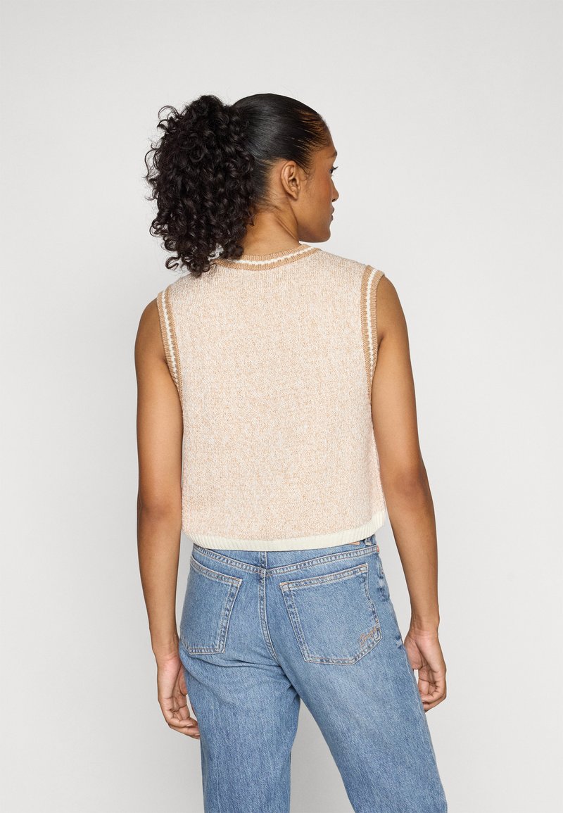 Ärmelloses Strickoberteil in hellbeige mit gerippten Kanten, kombiniert mit hoch geschnittenen blauen Jeans. Die Textur wirkt weich und leicht flauschig.