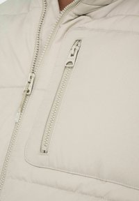 Lys beige puffjakke med en struktureret stof, der har en frontlynlås og en brystlomme med lynlås, der fremhæver praktiske designelementer.