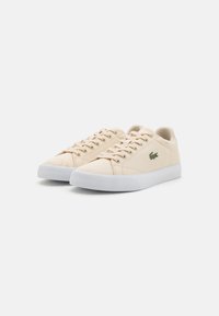 Beige lerret sneakers med hvit gummisåle, med snøring og en liten grønn krokodillelogo på siden.