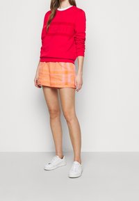 Röd sweatshirt med text, matchad med orange mönstrade shorts som har geometriska mönster, och vita sneakers. Mjuk tyg med avslappnad passform.
