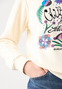 Sweter w kolorze kremowym z kolorową florystyczną i geometryczną grafiką. Miękka tkanina z prążkowanymi mankietami. Noszony z dżinsami.