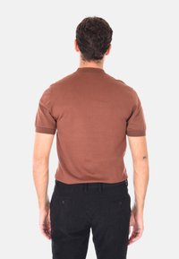 Uomo con capelli scuri e corti, indossa una camicia marrone a maniche corte e pantaloni neri, in piedi con le spalle rivolte verso l'osservatore su uno sfondo leggero e uniforme.