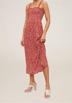 Woman wearing a sleeveless red midi dress with white floral print and white open-toe heeled sandals, standing against a plain background.