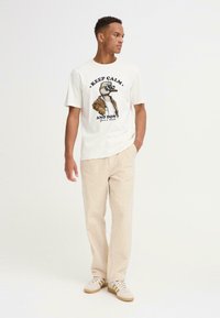 Hvid bomulds T-shirt med en tegneserieand-graphic iført en brun jakke, tekst "BEHOLD ROEN OG LAD VÆRE MED", parret med beige kordfløjl bukser og sneakers.