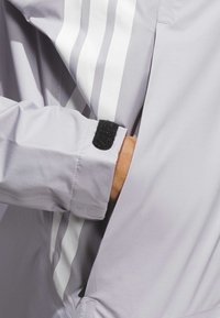 Light gray jacket with white stripes, featuring a Velcro adjustable cuff and a side pocket. Smooth texture and breathable fabric.