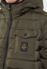 Giacca imbottita verde oliva con zip frontale, cappuccio, grande tasca sul petto e patch con logo. Presenta sezioni trapuntate per l'isolamento.