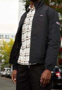 Person som bär en svart jacka över en beigefärgad rutig skjorta med knappar, stående utomhus med suddiga bilar och träd i bakgrunden.