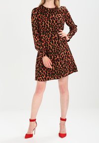 Robe à imprimé léopard avec de longues manches, encolure ronde et jupe évasée. Le tissu a une texture douce. Associée à des chaussures à talons rouges.