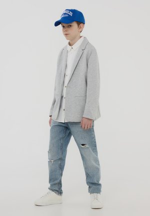 Blazer grigio sopra una camicia bianca, abbinato a jeans chiari strappati e sneakers bianche. Un cappellino blu completa l'outfit.