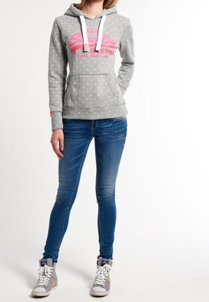 Sudadera gris con lunares, logo rosa, cordones blancos, bolsillo frontal, combinada con vaqueros azules y zapatillas altas grises.