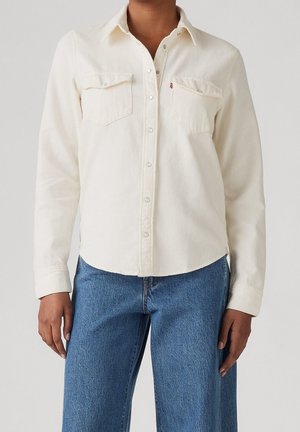 Person trägt ein langärmeliges, cremefarbenes Hemd mit Brusttaschen und blaue Jeans vor neutralem Hintergrund.