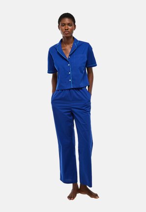 Femme debout pieds nus portant un haut bleu à manches courtes boutonné et un pantalon ample assorti sur un fond uni.