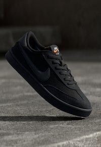 Baskets Nike noires avec une tige en daim et cuir, des lacets plats et une semelle texturée. Logo orange et jaune sur la languette.