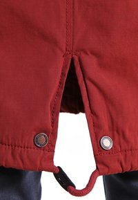 Ourlet de veste en tissu rouge avec une fente centrale, deux boutons-pression métalliques de chaque côté, et un cordon rouge avec une attache en plastique noir.
