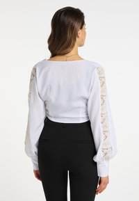 Weiße Bluse mit strukturierten Spitzeakzenten an den langen Ärmeln, tailliertem Bund und geknöpften Manschetten, kombiniert mit schwarzer, eleganter Hose.
