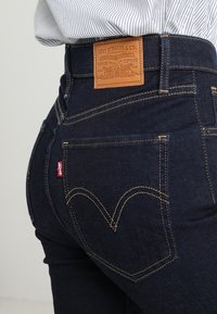 Tmavě modré džíny s vysokým pasem, se značením z kůže s vyraženým nápisem "Levi Strauss & Co." a žlutým prošíváním na zadních kapsách.