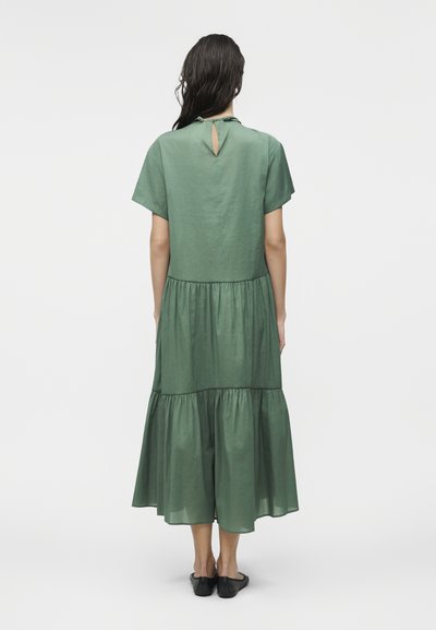 Γυναίκα που στέκεται γυρισμένη από την πλάτη, φορώντας ένα πράσινο φόρεμα μεσαίου μήκους με στρώσεις, με κοντά μανίκια και μαύρα φλατ παπούτσια.