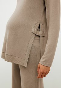 Beige Umstandsoberteil mit geripptem Saum und seitlichem Bindeband, aus weichem Stoff gefertigt, mit einem lockeren Schnitt, der einen wachsenden Bauch bequem umschließt.