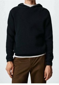 Hoodie noir en tricot avec un motif texturé, comprenant un col en V et des poignets côtelés. Porté sur une chemise blanche et associé à un pantalon marron.