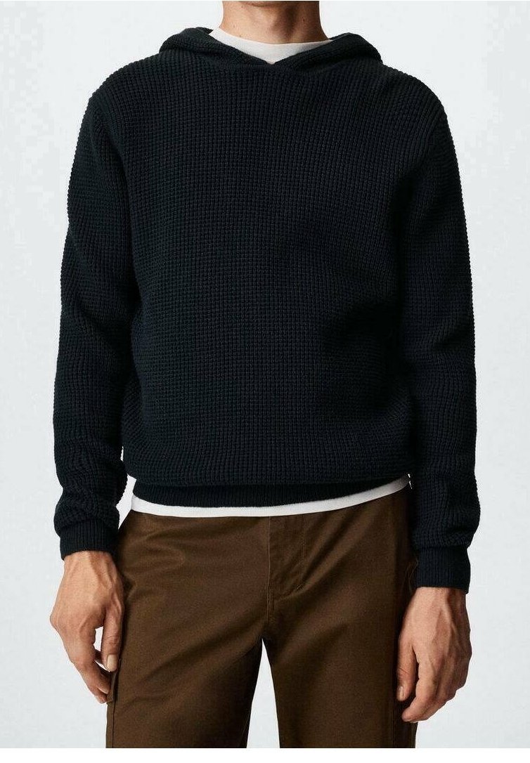 Hoodie noir en tricot avec un motif texturé, comprenant un col en V et des poignets côtelés. Porté sur une chemise blanche et associé à un pantalon marron.