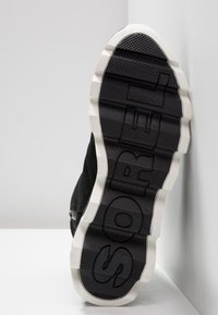 Sporta apavi ar melnu zamšu augšdaļu un biezu, baltu gumijas zoli, kurai ir teksturēti raksti un uz kuras apakšas iepresēts "SOREL".