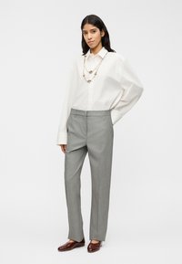 Ανοιχτό γκρι παντελόνι ραμμένο με λεία υφή, συνδυασμένο με λευκό πουκάμισο σε oversized γραμμή. Αξεσουάρ περιλαμβάνουν αλυσίδα για το λαιμό και καφέ παπούτσια.