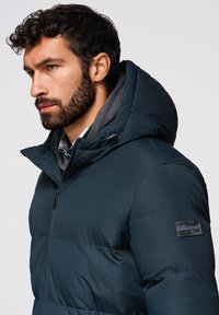 Middengrijze pufferjack met een afneembare capuchon, een ritssluiting aan de voorkant en een gestructureerd oppervlak; heeft een logo patch op de mouw.