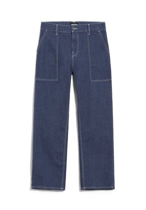 Žena v bílém tričku, černém svetru zavázaném kolem ramen a tmavě modrých širokých kalhotách s vysokým pasem, bosá na světlé podlaze.