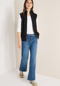 Černá pletená vesta na zip, nošená přes bílou košili s dlouhým rukávem a doplněná světle modrými džínami s širokými nohavicemi a béžovými teniskami.