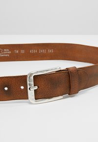 Bruinleren riem met een gestructureerde afwerking, zilveren metalen gesp en meerdere verstelgaten. Merken geven de maat en de oorsprong van het materiaal aan.