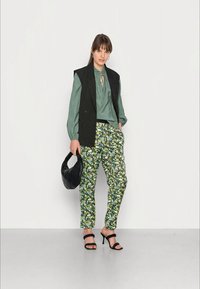 Colete riscas pretas sobre uma blusa verde, calças com padrão floral, sapatos de salto pretos com tiras e uma bolsa preta texturizada.