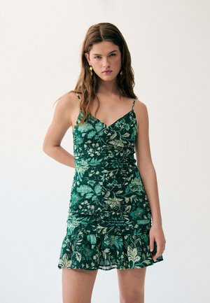Femme aux longs cheveux bruns portant une robe fleurie vert foncé sans manches avec un ourlet volanté, debout devant un fond clair uni.