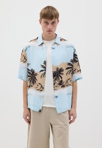 Camisa de manga corta en azul claro, con un estampado de playa y palmera en negro y beige. El modelo lleva una camiseta blanca debajo.