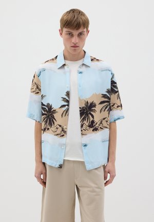 Chemise à manches courtes bleu clair, avec un motif de plage et de palmier en noir et beige. Le modèle porte un t-shirt blanc en dessous.