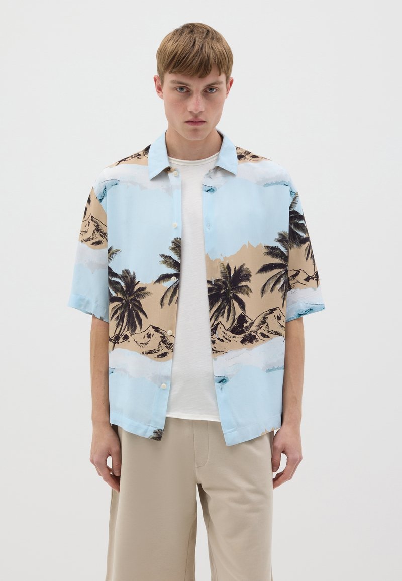 Camisa de manga corta en azul claro, con un estampado de playa y palmera en negro y beige. El modelo lleva una camiseta blanca debajo.