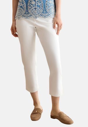 Mujer con pantalones blancos cortos, top azul de encaje y mocasines marrones sin cordones, de pie con una mano apoyada a un lado.