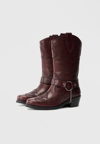 Vīna krāsas ādas kovboju zābaki ar izsmalcinātu šuvumu, ass purngalu un apaļu papēdi. Aprīkoti ar siksnu ar apaļu metāla detaļu.