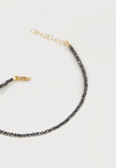 Λεπτό βραχιόλι με μικρές μαύρες βαθμιδωτές χάντρες και ρυθμιζόμενο κούμπωμα αλυσίδας σε χρυσό χρώμα σε απλό ανοιχτό φόντο.