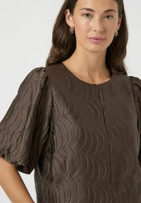 Brun, tekstureret top med en bølgeformet mønster, korte puff ærmer, rund halsudskæring og subtil glans. Minimale knapper i midten.