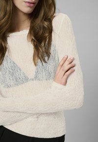 Off-white lace long-sleeve top with a sheer design, featuring a blue patterned underlayer. Smooth texture, crew neck, and fitted sleeves.